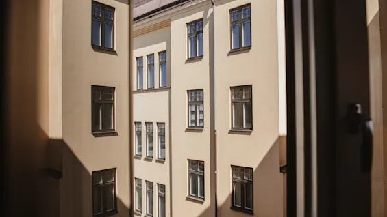 En beige bygning med flere vinduer ses gennem et åbent vindue. Sollys kaster skygger på facaden. Scenen er indrammet af vinduets kant.