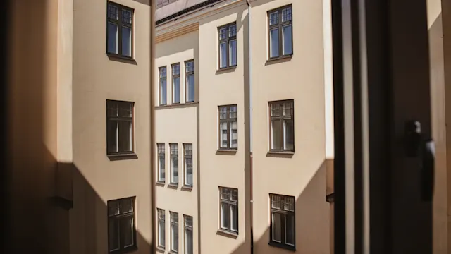 En beige byggnad med flera fönster ses genom ett öppet fönster. Solljus kastar skuggor på fasaden. Scenen ramas in av fönsterkanten.