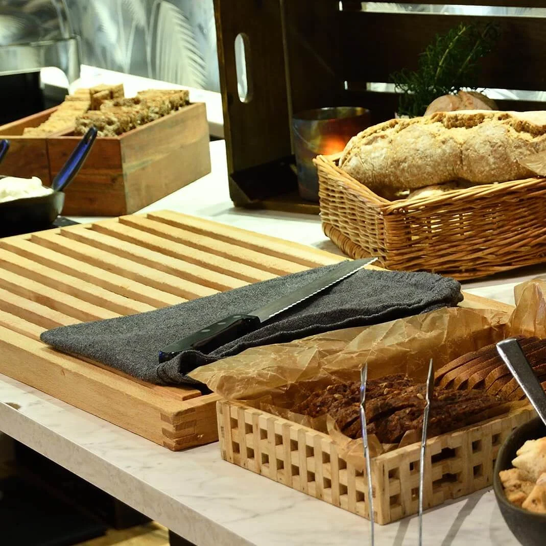 Puinen leikkuulauta, jossa on veitsi, ympäröitynä erilaisilla leipä- ja rullakorilla. Kulhot sisältävät siemeniä ja krutonkeja; suuri musta kattila viittaa buffet-tilanteeseen.