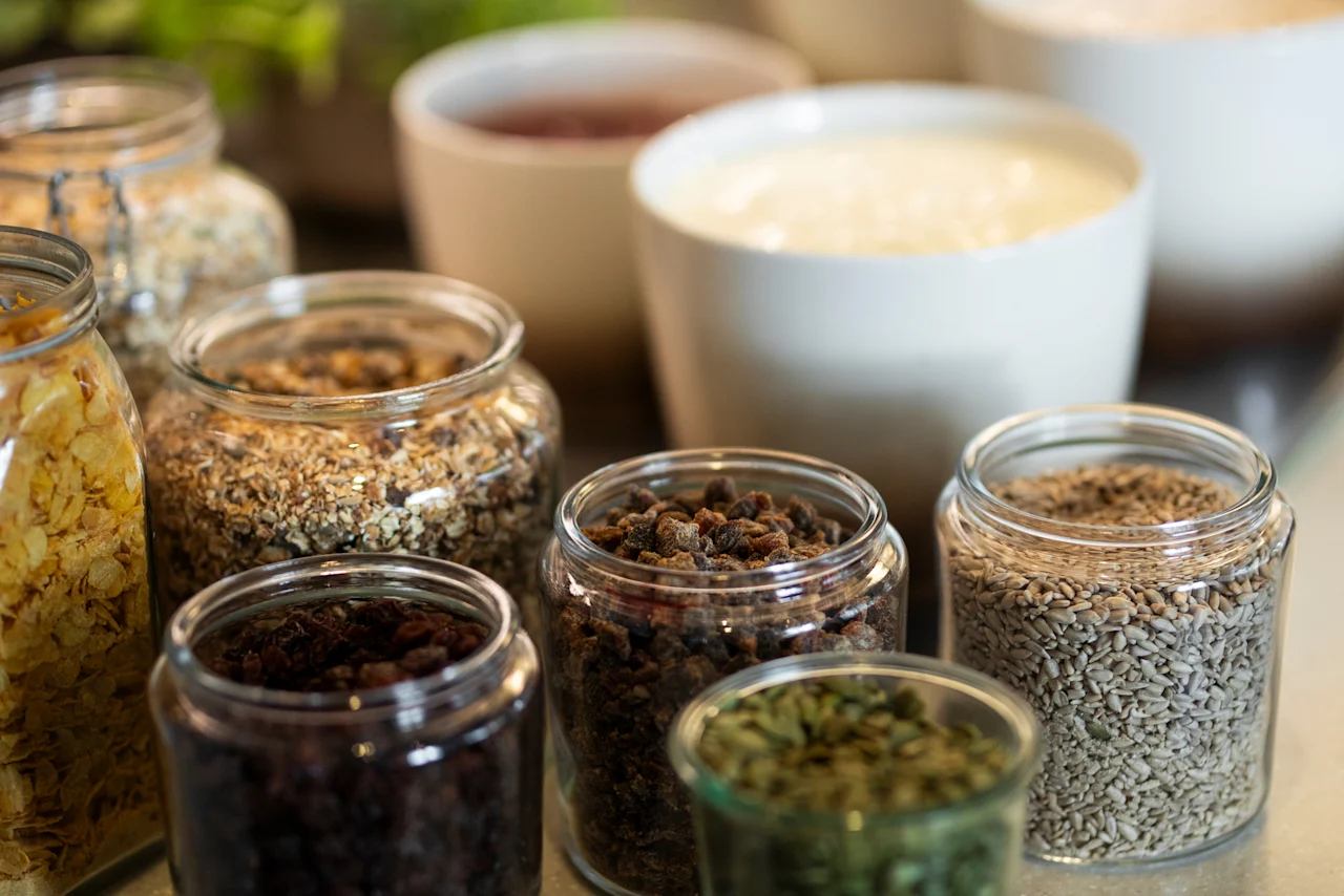 Ett urval av frukostingredienser i glasburkar och skålar, med olika flingor, torkad frukt och krämig yoghurt.