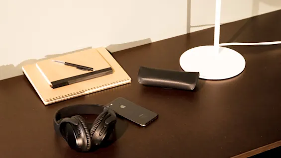 Headphones rest on a dark desk, accompanied by a smartphone, a closed notebook with pens, a glasses case, and a white desk lamp. Brown chair visible beneath.