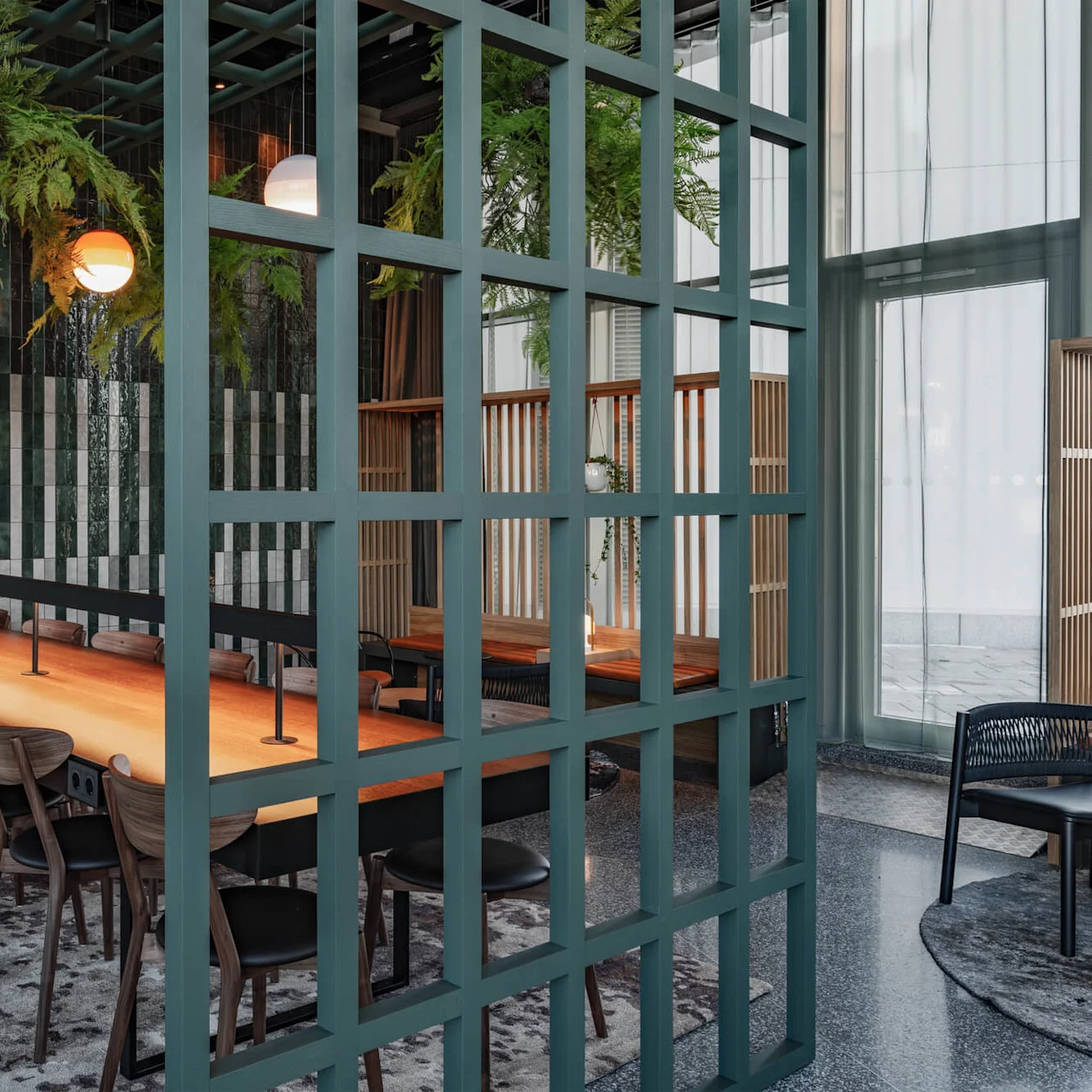 A long wooden table with chairs sits beneath hanging plants and round lights in a modern room with green tile walls, a grid partition, and large windows.