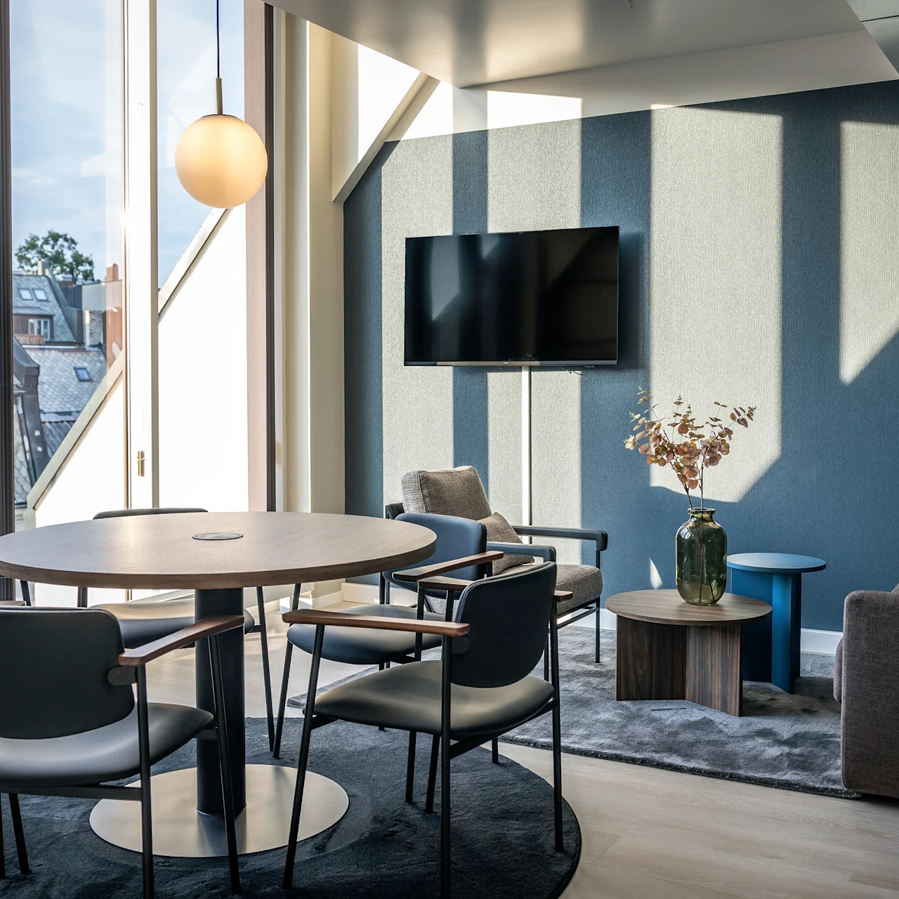Runt träbord omgivet av fyra stolar, upplyst av hängande lampa; beläget nära stora fönster med takutsikt och intill en loungedel med soffa, TV och dekorativt bord.