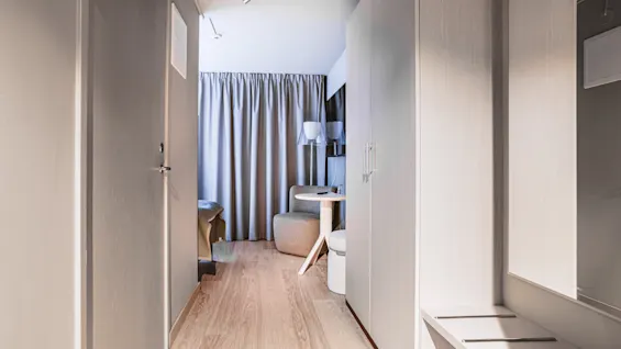 A bright, modern hotel room entrance with a hallway leading to a seating area with a table and curtains.