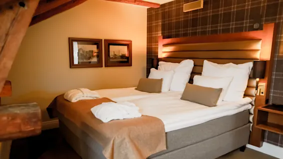A neatly made double bed with white linens and a tan throw in a cozy bedroom with warm lighting and framed pictures.