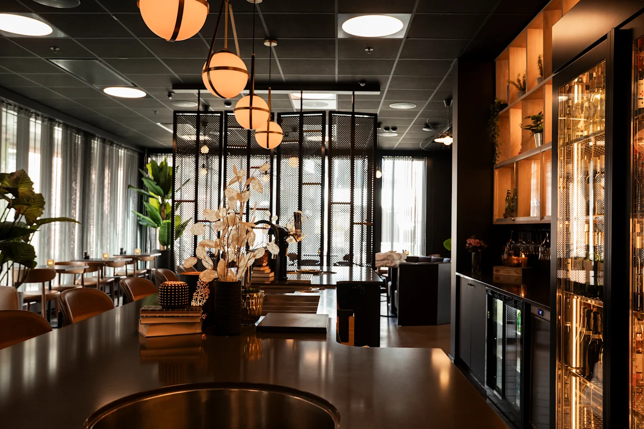 Et elegant, dempet opplyst bar- eller restaurantinteriør med varm belysning, en lang disk og sitteplasser i bakgrunnen.
