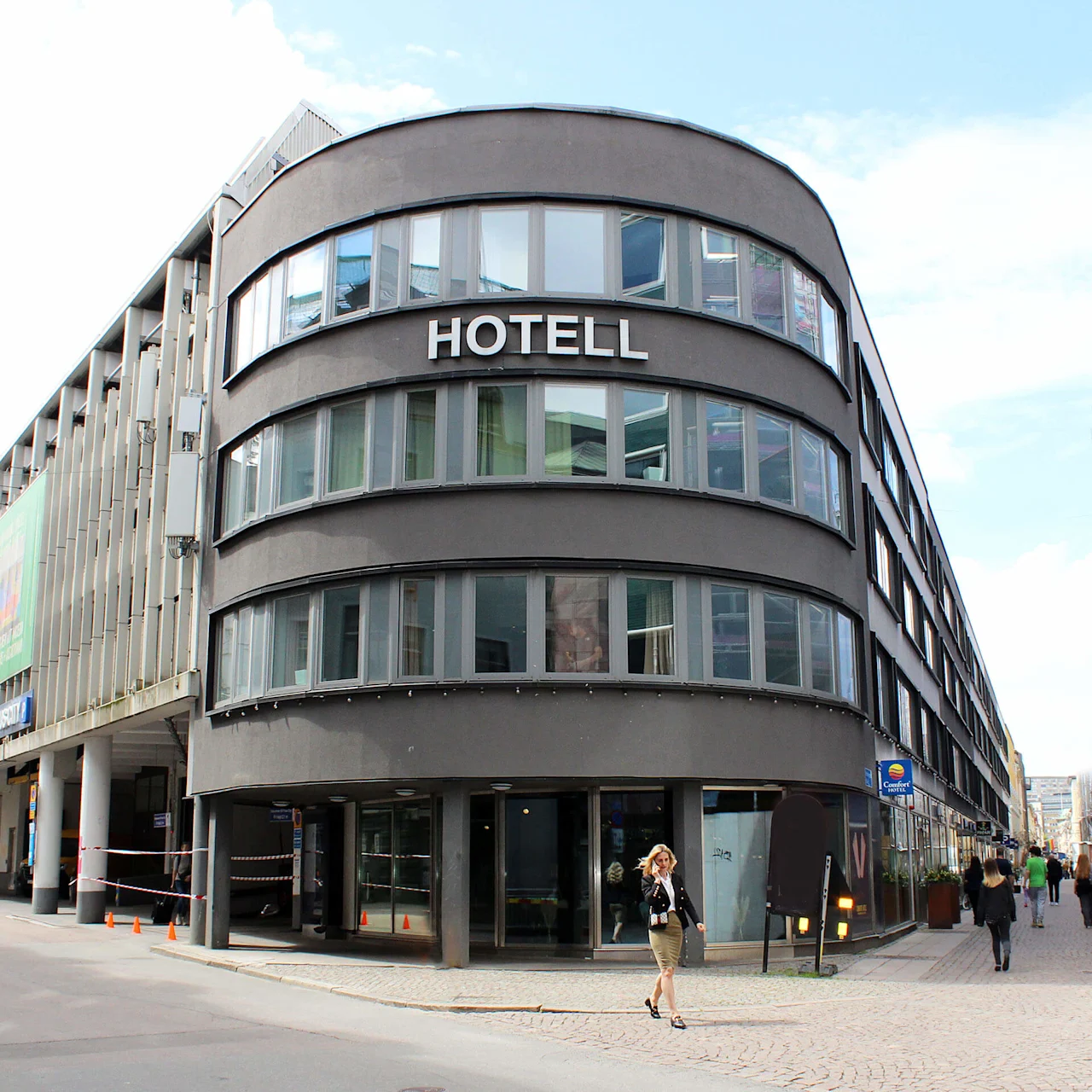 En buet moderne bygning merket "HOTELL" med store vinduer; en person går på brostein ute. Omkringliggende bygninger og en klar himmel gir en urban atmosfære.