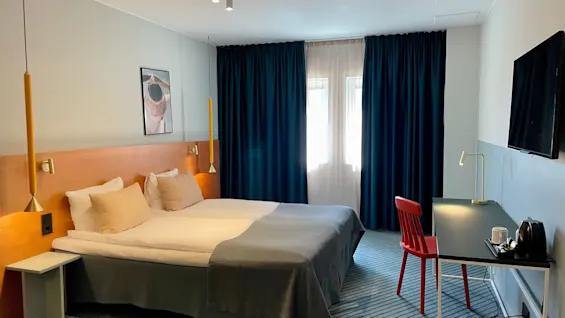 En pent oppredd seng står i et moderne hotellrom; den vendes mot en veggmontert TV. Et skrivebord med en rød stol og lampe er i nærheten. Blå gardiner dekker vinduene.