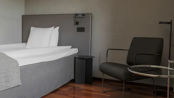 A neatly made bed with white pillows is adjacent to a black nightstand. Beside it, a modern black armchair and round glass table rest on a wooden floor.