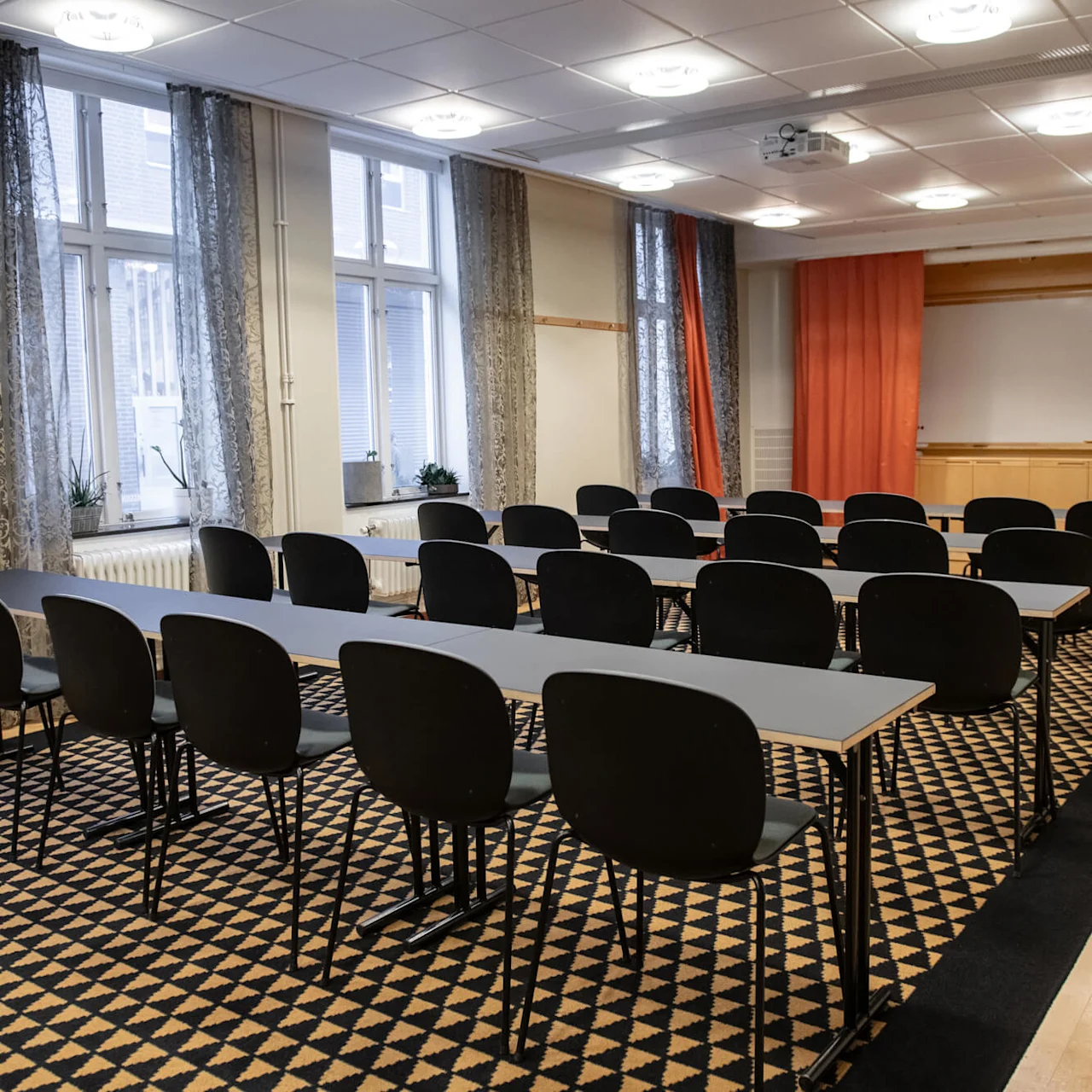 Rows of empty black chairs face a podium with a laptop in a conference room, featuring patterned carpet and large curtained windows.