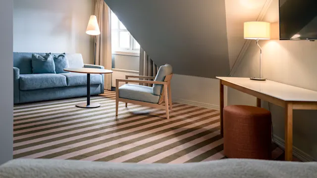 Et koselig stue med en blå sofa, lenestol og rundt bord. En gulvlampe og TV pryder rommet med skråtak og stripete teppe.
