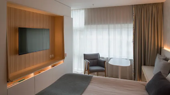 A wall-mounted TV hangs in a wood-paneled nook beside a bed with gray bedding. A chair and small table sit by a large, softly-lit window.