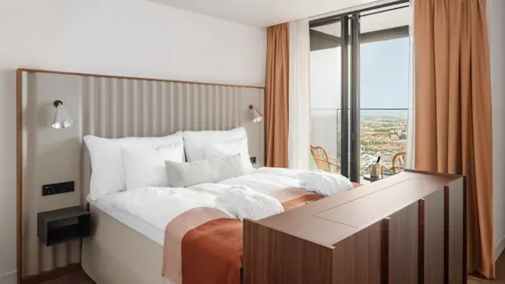 En prydligt bäddad säng med vita kuddar och en rostfärgad filt står i ett modernt hotellrum. Fönster från golv till tak avslöjar en stadsbild och två stolar utanför.
