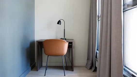 En brun stol står foran et træskrivebord med en sort lampe, i et hjørnerum med lyse vægge og store gardiner.