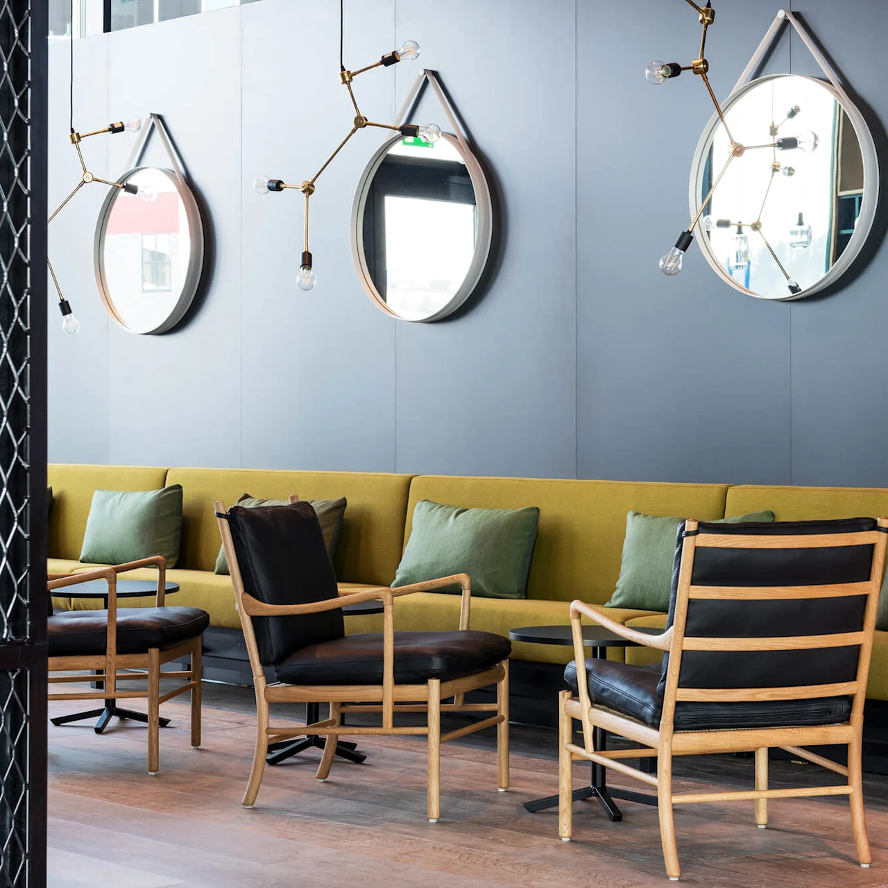 Elegant chairs in a lounge setting face a modern couch with green pillows, framed by blue walls adorned with mirrors and unique light fixtures. A patterned divider partially shows the space.