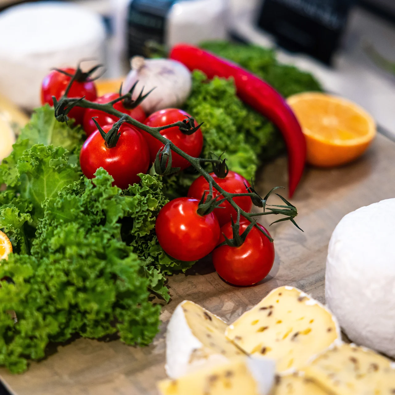 Kirsikkatomaatit lepäävät kihara salaattialustalla, ympärillään juustoa, valkosipulia, chilipippureita ja halkaistuja appelsiineja puupinnalla.