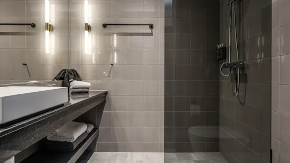 Moderne badeværelse med en hvid vask på en sort bordplade oplyst af to lodrette lys. Håndklæder og toiletartikler er pænt arrangeret med en bruseenhed i glas i nærheden.