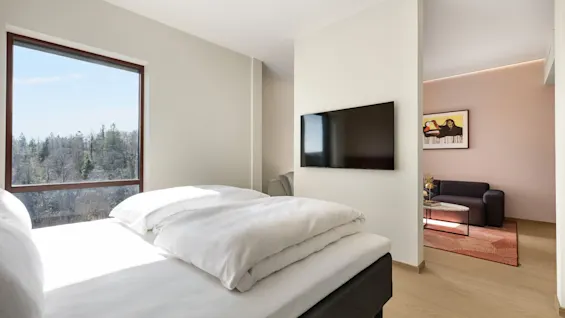 Et lyst hotellrom med en seng, et stort vindu med utsikt over en skog, og et separat salongområde med sofa og kunst.