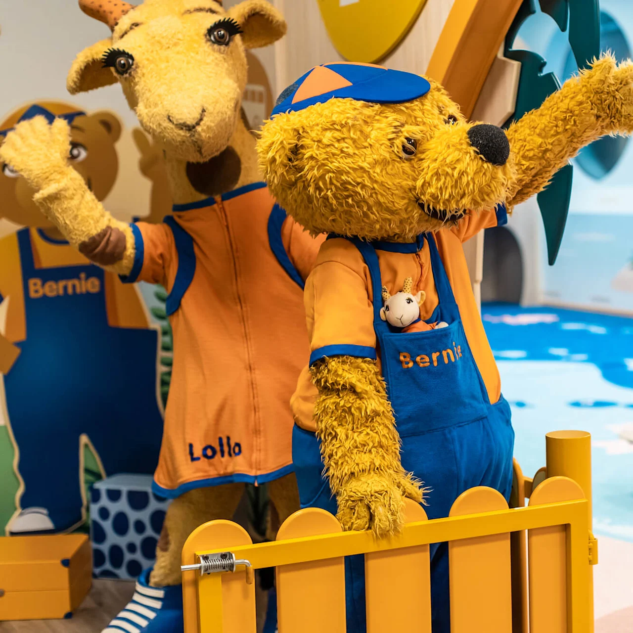 En bjørn iført orange og blå dragt med teksten "Bernie" vinker ved siden af en giraf mærket "Lollo" i et farverigt, legende indendørsmiljø med et legetøjs-surfbræt.