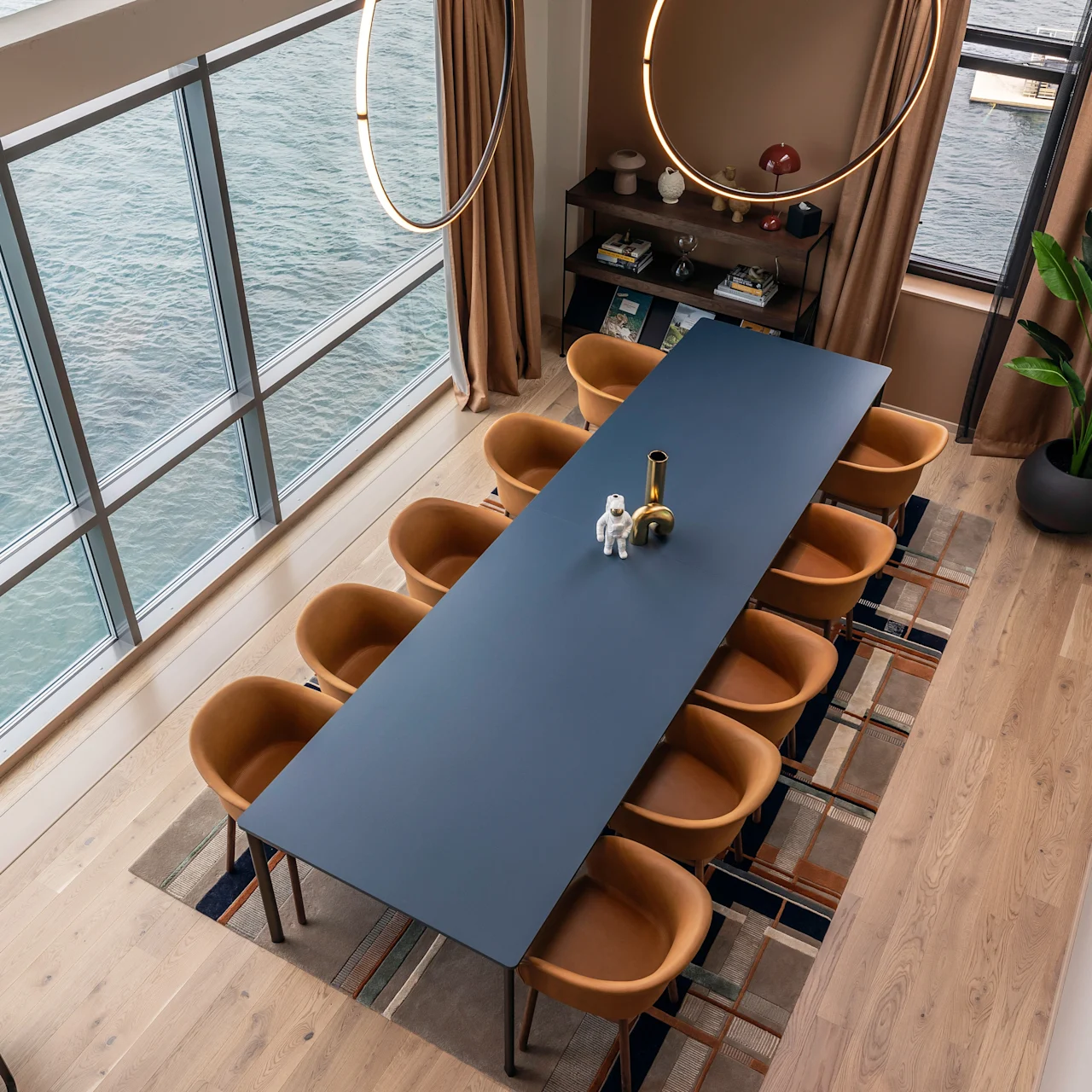 Langt bord med en vase og figur placeret i et moderne rum, omgivet af brune stole. Store vinduer viser udsigt over vandet, med indendørs planter og dekorative genstande, der forbedrer indretningen.