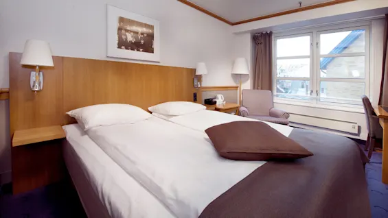 Et pænt hotelværelse med to enkeltsenge, en stribet sofa, og et arbejdsbord nær et vindue med udsigt til byen.