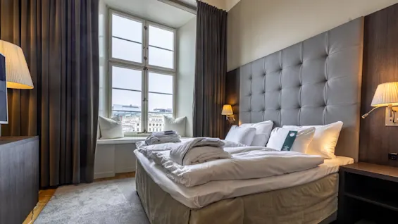 A neatly made bed with white linens and plush pillows rests under a tufted headboard, flanked by wall lamps in a room with large windows overlooking a cityscape.