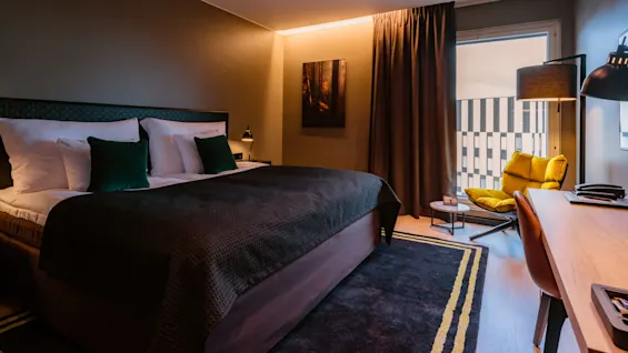 A bed with dark bedding and green cushions stands in a modern bedroom with neutral walls, a wooden desk, yellow chair, and large window with heavy curtains.
