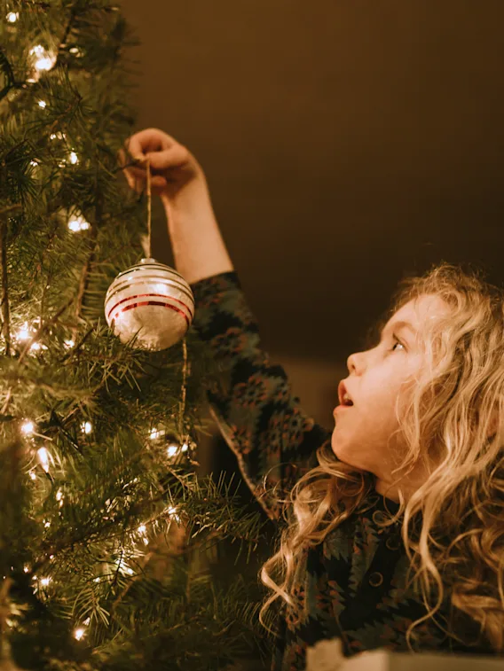 Ei liten jente med krøllete hår henger opp en julekule på et stemningsfullt, lyst juletre.