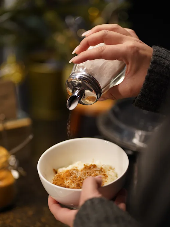En persons händer strör socker från en ströare över en skål med efterrätt med kanel.
