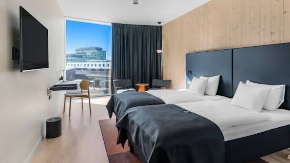 Et lyst, moderne hotelværelse med to senge, stort vindue med byudsigt, skrivebord og vægmonteret TV.
