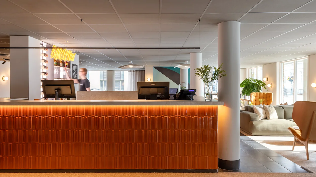 Reception med datorer och en person bakom. Neonljusskylt lyder "BOKA RUM." Modern lounge med soffor och växter. Stora fönster släpper in naturligt ljus.