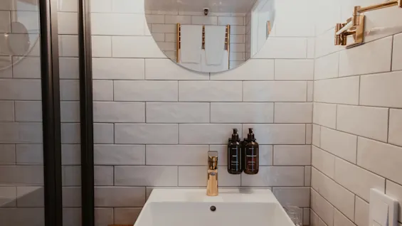 Et moderne badeværelse med hvide flisevægge, en hvid vask, guldkran og sæbe- og body lotion-dispensere. Et rundt spejl hænger ovenover.