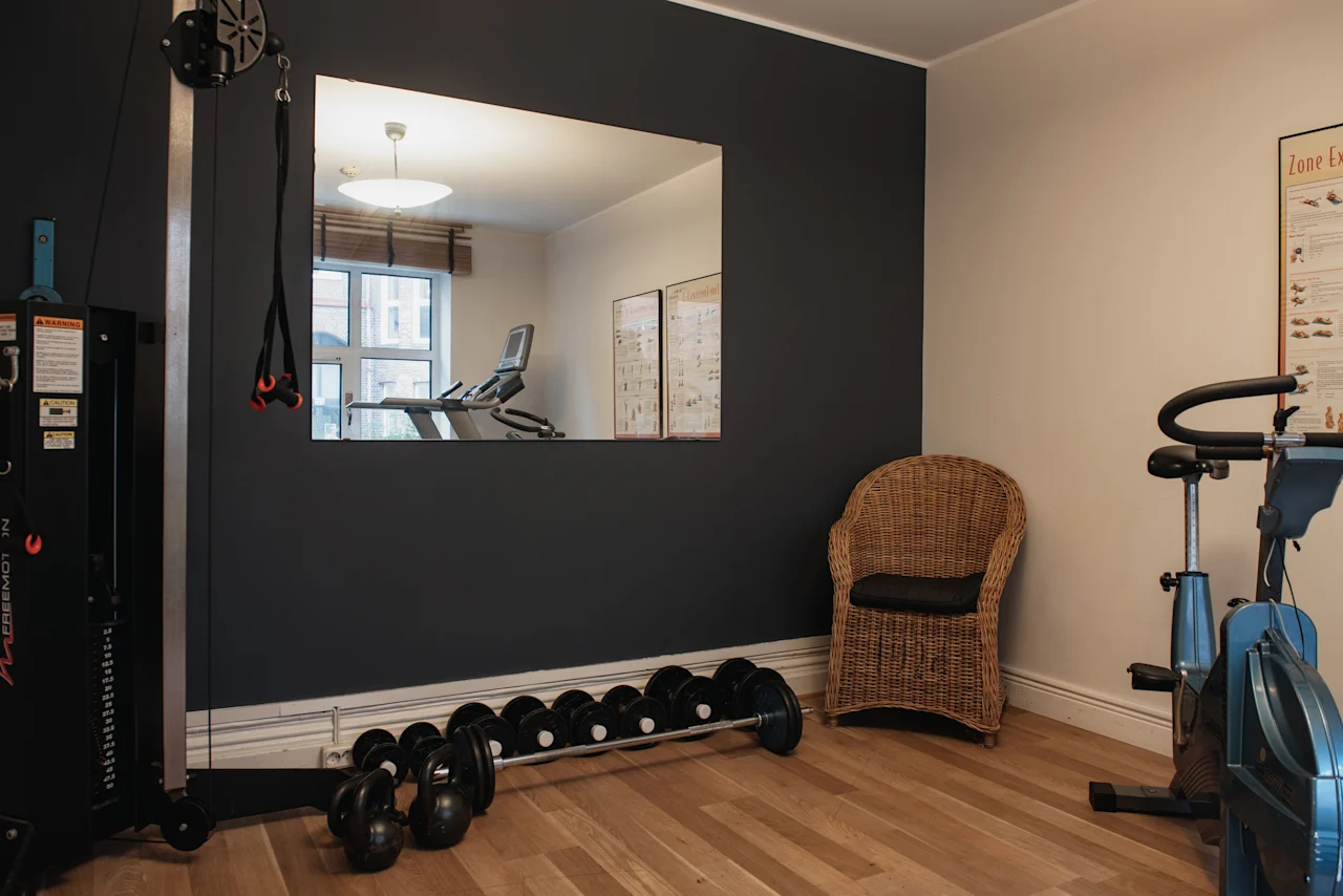Et fitnessrum med en vægtmaskine, håndvægte, kettlebells, en motionscykel og et spejl, der reflekterer et løbebånd. Zone Ex-plakat.