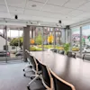 Et langt konferansebord med stoler er plassert i et moderne møterom. Store vinduer viser en gate utenfor med parkerte biler og en fargerik veggmaleri på bygningen.