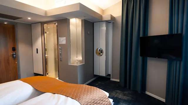 Hyvin valaistu hotellihuone, jossa kaksi siististi pedattua sänkyä, oranssi peitto ja verhot. Taulutelevisio on asennettu seinälle. Näkyvissä on lasisuihku ja puuovi.