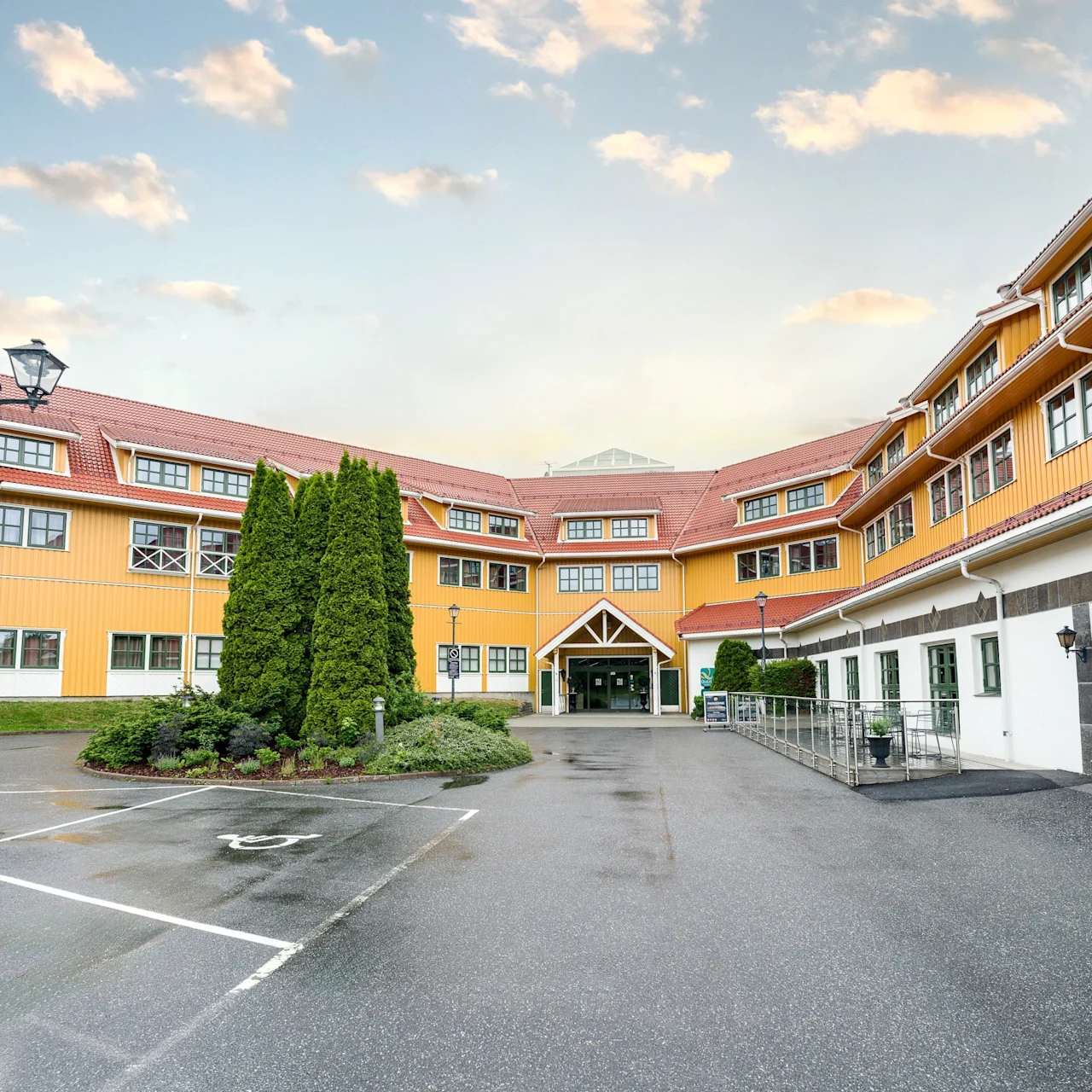 A large yellow building with red roofs and white-trimmed windows stands prominently. A ramp and entrance sign labeled "Social" are seen. Tall trees line the foreground. Cloudy sky above.