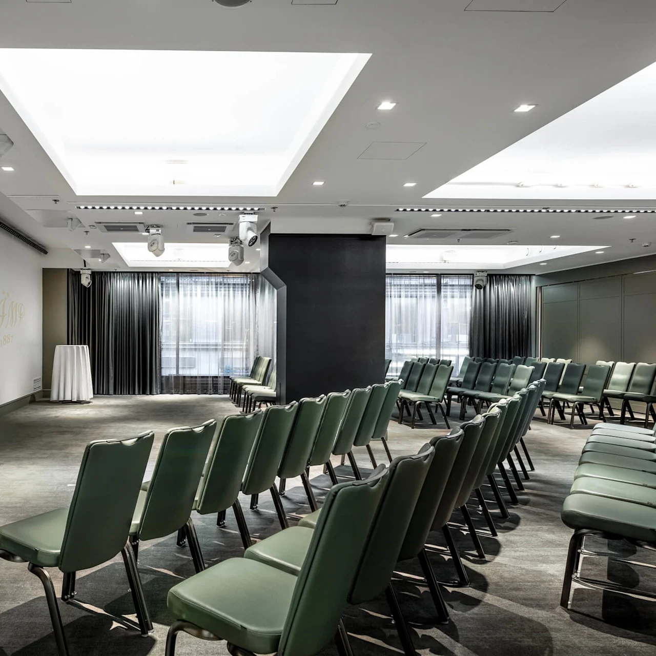 Rows of green chairs face a wall with "KÄMP 1887" written on it, in a well-lit conference room with ceiling lights and curtains.