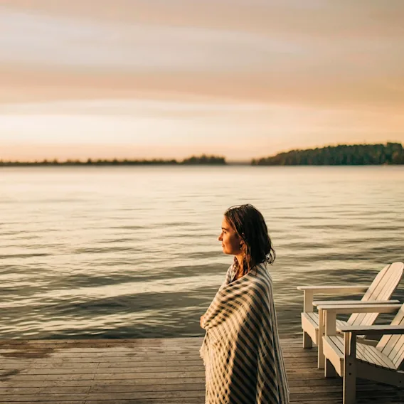Person wrapped in a blanket on a dock, gazing at a serene lake during a beautiful sunset.