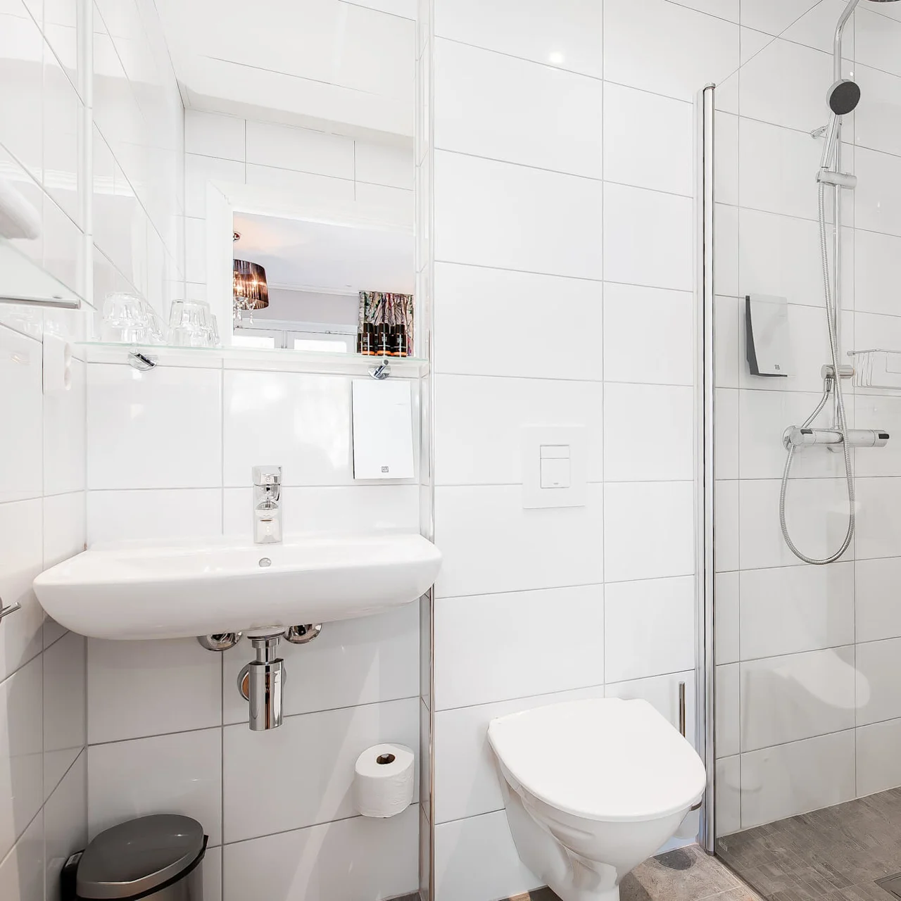 Et moderne bad med toalett, vask og dusj inngjerdet med glass, hvite fliser og reflekterende overflater.