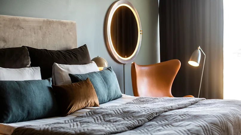 Bed adorned with multi-colored pillows, covered by a quilt, positioned near a brown chair under a mirror with lights and beside a lamp; set in a softly-lit, modern room.