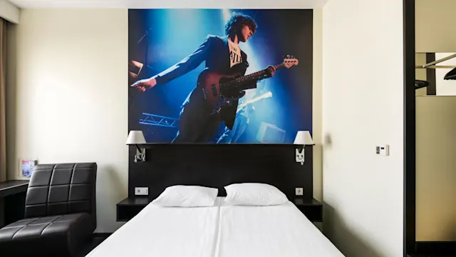 Bed with white linens, positioned below a large mural of a musician playing a guitar on stage. Room includes a dark chair and a small table by the window.