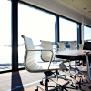 A modern conference room with white chairs and a large table, sunlight streaming in, and a sea view through floor-to-ceiling windows.