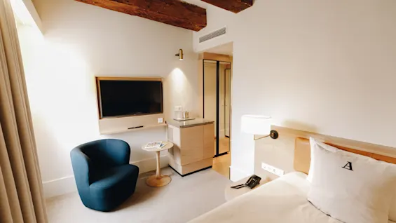 Ett mysigt hotellrum med en bekväm säng, en blå fåtölj och en väggmonterad TV, med rustika träbjälkar.