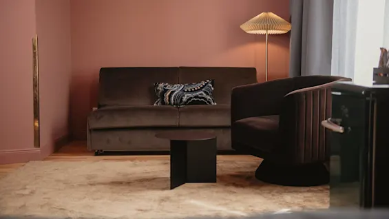 En brun soffa med dekorativa kuddar står mot en rosa vägg, med en stilren lampa och fåtölj bredvid ett litet mörkt bord i ett mysigt rum.