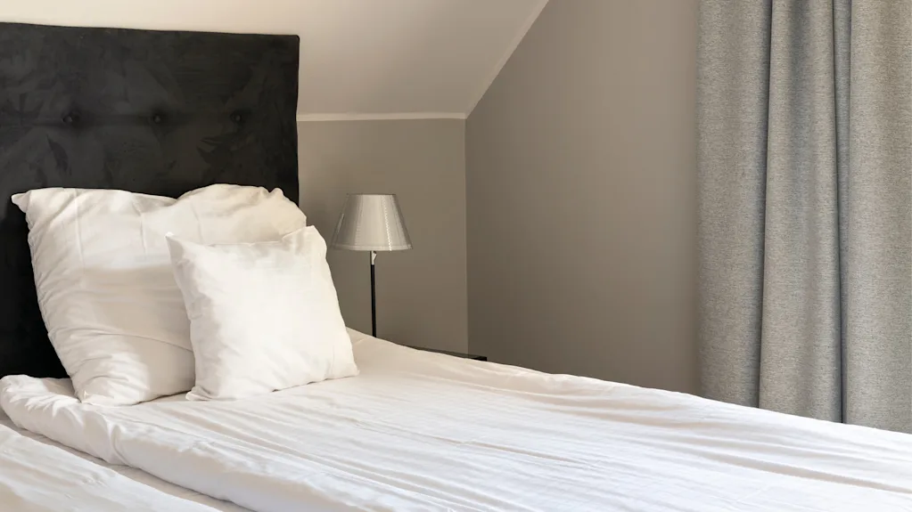 Bed with white linens and two pillows rests against a dark headboard. A small lamp stands on a bedside table, with grey curtains drawn in a softly lit bedroom.