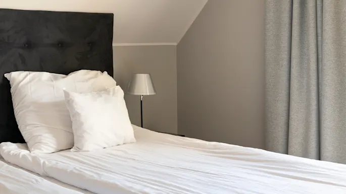 Seng med hvitt sengetøy og to puter hviler mot en mørk sengegavl. En liten lampe står på et nattbord, med grå gardiner trukket for i et mykt opplyst soverom.