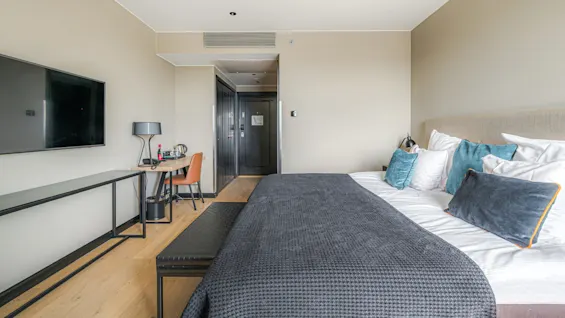 A neatly made bed with multiple pillows and a dark coverlet sits in a modern hotel room. A wall-mounted TV, desk with a chair, and lamp complete the minimalistic decor.