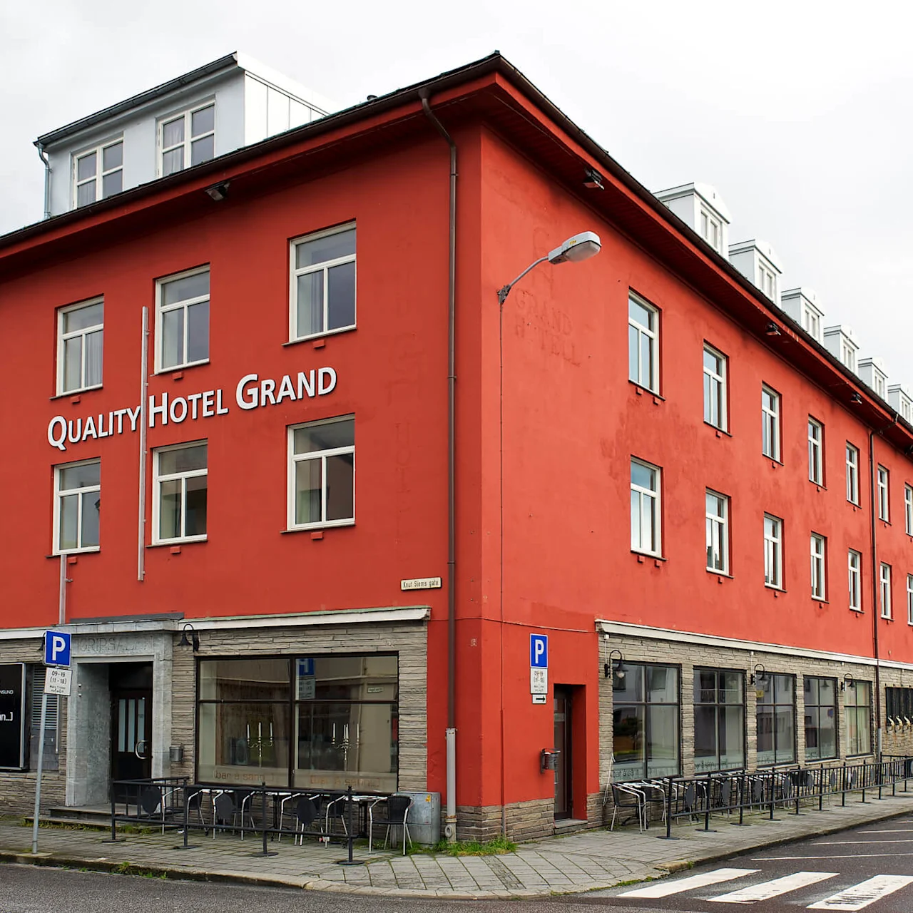 En rød bygård med flere etasjer, merket "QUALITY HOTEL GRAND", står på et hjørne. Fortauet har parkering forbudt-skilt og uteservering. Overskyet vær med omkringliggende bygninger synlige.