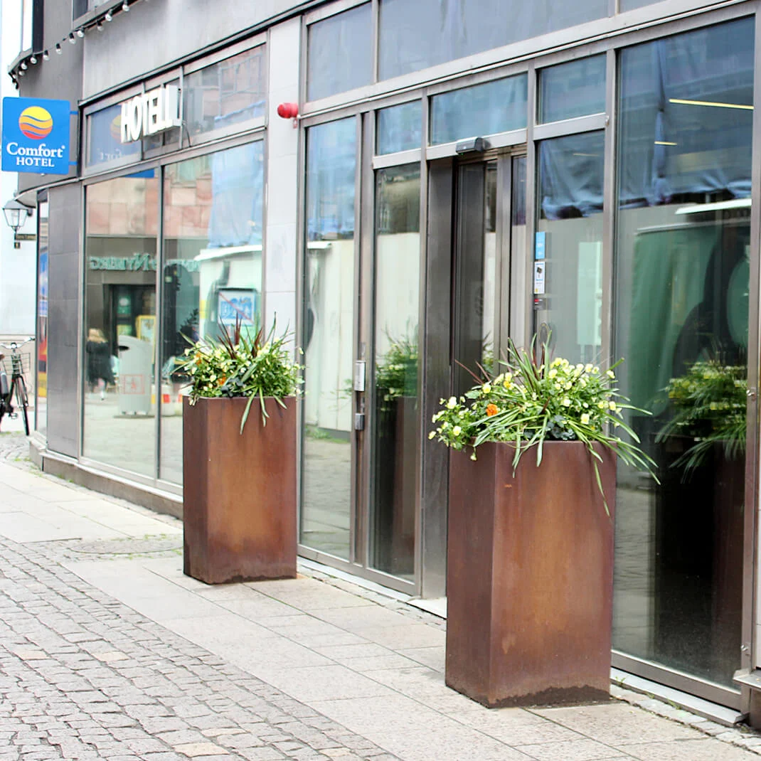 To stiliserte krus danner ansikter; ett har et dampende hjerte over. "COMFORT HOTEL - FOR URBAN EASY LIVING" står skrevet. Det er ute, med planter og brostein.
