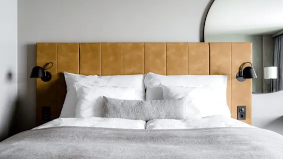A neatly made bed with white pillows and a beige blanket is centered against a brown upholstered headboard. Black wall lamps are mounted on each side, with a large mirror partly visible.
